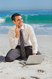 Thoughtful businessman sitting on the sand with his laptop