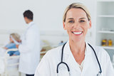 Smiling blond doctor posing with doctor attending patient on background