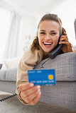 Happy young woman with credit card laying on sofa and talking ph