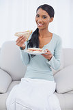 Smiling attractive woman sitting on cosy sofa eating sandwich