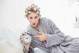 Astonished blonde woman in hair curlers pointing her alarm clock