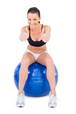 Positive woman in sportswear sitting on exercise ball giving thumbs up to camera