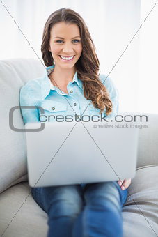 Cheerful brunette sitting on sofa using laptop
