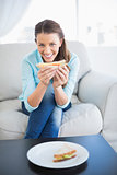 Happy woman sitting on sofa showing sandwich at camera
