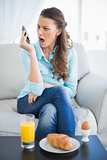 Angry woman screaming on the phone sitting on sofa