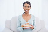 Smiling attractive woman sitting on cosy sofa holding sandwich