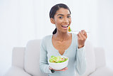 Smiling attractive woman sitting on cosy sofa eating salad