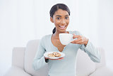 Smiling attractive woman sitting on cosy sofa holding coffee and cookies