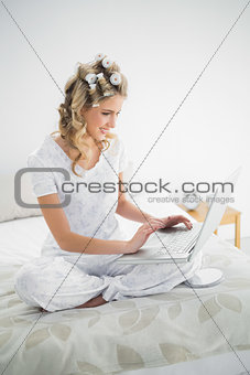 Smiling cute blonde wearing hair curlers using laptop