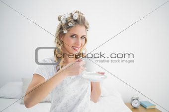 Relaxed pretty blonde wearing hair curlers holding coffee
