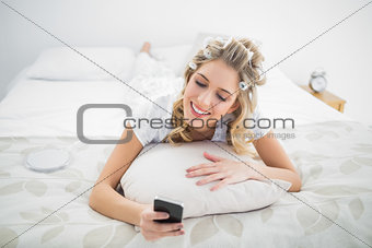 Peaceful blonde wearing hair curlers texting while lying on bed