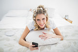 Smiling blonde wearing hair curlers holding smartphone while lying on bed
