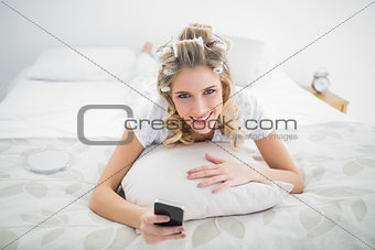 Smiling blonde wearing hair curlers holding smartphone while lying on bed