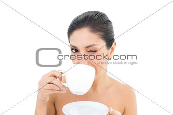Pretty brunette holding a cup and saucer and taking a sip while winking