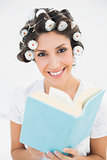 Smiling brunette in hair rollers holding a book on bed