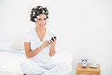 Happy brunette in hair rollers sending a text on bed