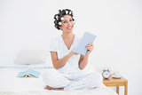 Pretty brunette in hair rollers using tablet on bed smiling at camera