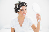 Happy brunette in hair rollers holding hand mirror smiling at camera
