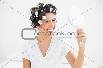 Happy brunette in hair rollers holding hand mirror smiling at camera