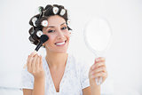 Smiling brunette in hair rollers holding hand mirror and applying makeup