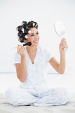 Cheerful brunette in hair rollers holding hand mirror and applying makeup