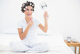 Cheerful brunette in hair rollers holding hand mirror and applying lip gloss