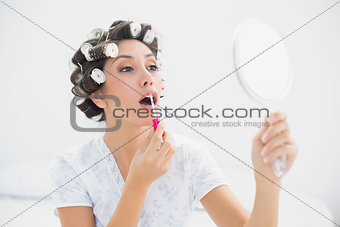 Pretty brunette in hair rollers holding hand mirror and applying lip gloss