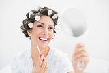 Happy brunette in hair rollers looking in hand mirror and holding  lip gloss
