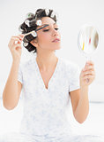Happy brunette in hair rollers looking in hand mirror and brushing her eyebrows