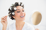 Smiling brunette in hair rollers looking in hand mirror and brushing her eyebrows