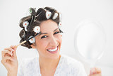 Pretty brunette in hair rollers holding hand mirror and brushing her eyebrows