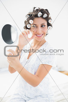 Pretty brunette in hair rollers looking in hand mirror and using eyelash curler
