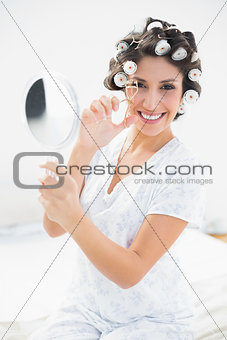 Pretty brunette in hair rollers holding hand mirror and using eyelash curler