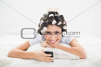 Happy brunette in hair rollers lying on her bed sending a text