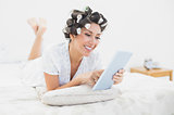 Happy brunette in hair rollers lying on her bed using her tablet