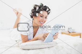 Happy brunette in hair rollers lying on her bed using her tablet