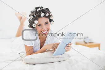 Smiling brunette in hair rollers lying on her bed using her tablet