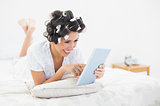 Cheerful brunette in hair rollers lying on her bed using her tablet