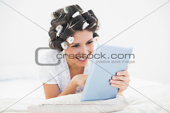 Pretty brunette in hair rollers lying on her bed using her tablet