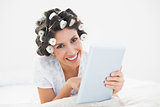 Pretty brunette in hair rollers lying on her bed using her tablet smiling at camera