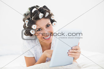 Pretty brunette in hair rollers lying on her bed using her tablet smiling at camera