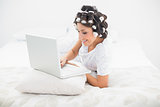 Pretty brunette in hair rollers lying on her bed using her laptop