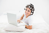 Happy brunette in hair rollers lying on her bed using her laptop