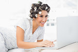Smiling brunette in hair rollers lying on her bed using her laptop