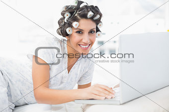 Cheerful brunette in hair rollers lying on her bed using her laptop