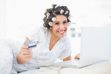 Cheerful brunette in hair rollers lying on her bed using her laptop to shop online