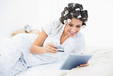 Excited brunette in hair rollers lying on her bed using her tablet to shop online