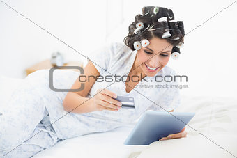 Excited brunette in hair rollers lying on her bed using her tablet to shop online
