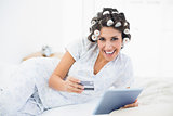 Happy brunette in hair rollers lying on her bed using her tablet to shop online