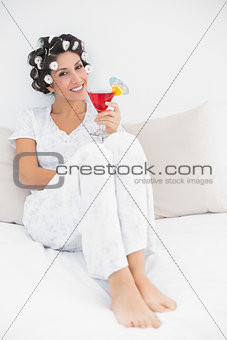 Cheerful brunette in hair curlers sitting on her bed holding a cocktail